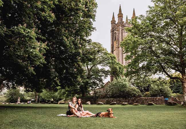 Two people having a picnic with their dog in the green in front of the church in Widecombe-in-the-Moor in Devon